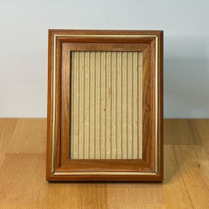 May include: A rectangular wooden picture frame with a medium brown finish and a thin gold-toned inner border. The frame contains a vertical piece of light tan corrugated cardboard as a backing, featuring a textured, ribbed surface pattern.