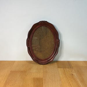 May include: An oval-shaped picture frame with a deep burgundy finish and ornate, raised floral detailing along the edges. The centre of the frame features a textured, brown, woven fabric insert, providing a contrast to the smooth, dark frame.