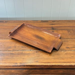 May include: A rectangular wooden serving tray in a warm brown hue. The tray has a gently curved design and includes built-in handles at either end. The natural wood grain is visible. Suitable for serving food or displaying decorative items. The tray's dimensions are approximately 40 cm in length.
