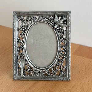 Puede incluir: Un marco de fotos rectangular plateado con una abertura ovalada. El marco está decorado con un diseño floral y de vid, y un pájaro está posado en la esquina superior derecha. Un cáliz está en la esquina inferior izquierda.