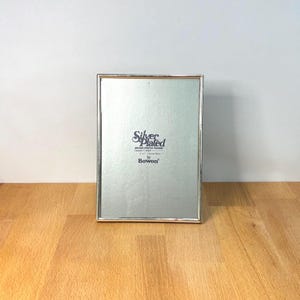May include: A rectangular silver-plated brass photo frame with a thin metallic border. The centre insert features the text Silver Plated Brass Photo Frame Lacquer Coated 5x7 13x18cm by Bowon in dark lettering against a light grey background.