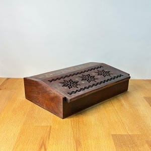 May include: A rectangular dark brown wooden box featuring a hinged lid with a decorative cutout pattern. The lid displays three prominent star-shaped motifs surrounded by a lattice design, with small circular inlays integrated into the wood surface.