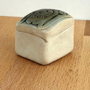 Könnte beinhalten: Eine quadratische Schmuckschatulle aus Keramik mit Deckel. Die Box ist hellbeige, der Deckel dunkelgrün mit einem wirbelnden, erhabenen Design. Die Box steht auf einer Holzoberfläche.