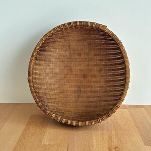 Pu&ograve; includere: Un cesto rotondo intrecciato di colore marrone naturale. Il cesto ha un design ampio e poco profondo con una base fitta e un bordo decorativo. Il bordo &egrave; rinforzato con una serie di supporti verticali. Il cesto &egrave; appoggiato su una superficie di legno.