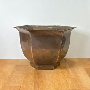 Op de afbeelding: Een zeshoekige koperen plantenbak met een gepolijste metalen afwerking en een uitlopende rand. De pot heeft een taps toelopend lichaam dat smaller wordt naar een platte, zeshoekige basis, met een warm, reflecterend oppervlak met kleurnuances.