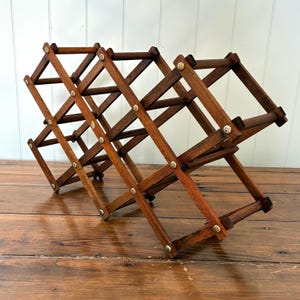 Pode incluir: Um suporte de vinho de madeira castanha escura com um design de treliça. O suporte dobrável é expansível. As juntas são fixadas com pequenos rebites metálicos decorativos. Projetado para conter várias garrafas de vinho.