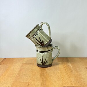 May include: Two ceramic mugs stacked vertically, featuring a speckled light green glaze with dark brown painted leaf-like motifs and a horizontal band. Each mug has a sturdy handle and a dark brown base, showcasing a handcrafted aesthetic.