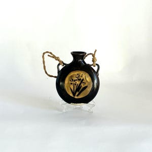 May include: A small, round, black ceramic flask with a narrow neck and two side handles. A circular gold-toned medallion on the front features a black botanical illustration of flowers. A piece of natural twine is tied through the handles.