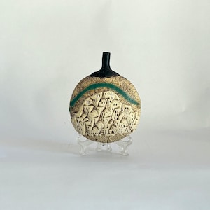 May include: A round, flat ceramic vase with a textured, beige surface featuring a relief carving of a hillside village. A smooth, dark brown neck extends from the top, and a curved teal band arches over the village. It rests on a clear stand.