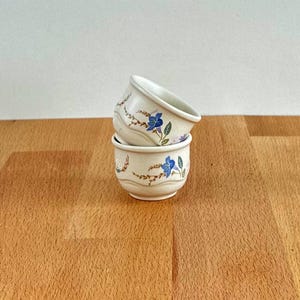 Könnte beinhalten: Zwei kleine, cremefarbene Keramikschalen, die übereinander gestapelt sind. Jede Schale hat ein Blumendesign mit blauen Blumen und grünen Blättern. Die Schalen stehen auf einer Holzoberfläche.