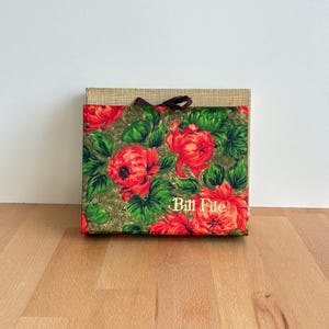 May include: A square box with a floral pattern of red flowers and green leaves on a gold background. The text "Bill File" is visible. A brown ribbon is tied around the box.
