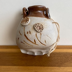 May include: A handmade ceramic vase with a speckled white body and a brown rim and handles. The vase features carved floral designs and two raised floral accents. A natural twine is attached for hanging.