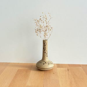 May include: A small, beige ceramic vase with a bulbous base and a long, slender neck. The vase has a brown, stylized leaf pattern around the base and neck. It holds a delicate arrangement of dried, light-coloured flowers. The vase sits on a wooden surface.