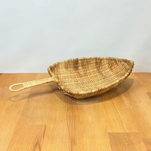 May include: A light brown woven wicker scoop with a long, slender handle. The scoop features a wide, open, leaf-shaped basket design constructed from natural reed material, showcasing a detailed, intricate, and uniform woven pattern throughout.