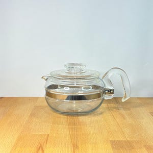 Könnte beinhalten: Eine Teekanne aus klarem Glas mit einem abgerundeten Körper und einem horizontalen, metallisch silbernen Band in der Mitte. Sie verfügt über einen passenden Glasdeckel mit einem kleinen Knauf und einen geschwungenen Glasgriff an der Seite.