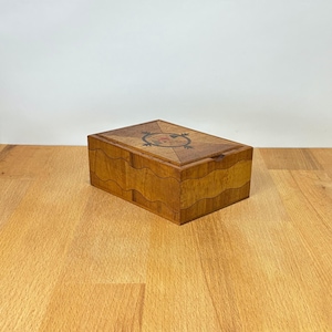 May include: A rectangular wooden box featuring a light and dark brown wood veneer finish. The lid displays a geometric starburst pattern with a central circular inlay. The sides of the box are constructed with a distinct wavy horizontal seam.