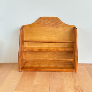 Puede incluir: Una estantería de madera para montar en la pared con tres niveles. La estantería está hecha de madera de color claro y tiene una parte superior curvada. Los estantes están soportados por piezas laterales verticales. La estantería está diseñada para exhibir objetos pequeños.