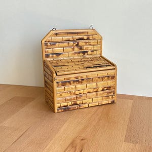 May include: A small, rectangular wooden box with a hinged lid and a backrest. The box is constructed from thin, rectangular pieces of wood, arranged to resemble bricks. The wood has a light brown color with darker, burnt-looking accents.