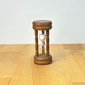 Puede incluir: Un reloj de arena con marco de madera marrón, compuesto por una base y una parte superior cilíndricas unidas por cuatro pilares verticales. La cámara central de cristal contiene arena blanca, parte de la cual descansa en el bulbo inferior.