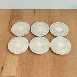 May include: Six silver-coloured coasters with an ornate, repeating floral pattern. The coasters are round and arranged on a wooden surface. Each coaster has a slightly raised edge and a detailed design.