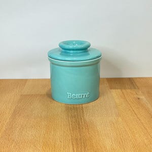 以下が含まれることがあります： 中央にノブが付いた蓋付きの円筒形ターコイズブルーの陶器製バターケース。容器の前面には筆記体でBeurreという文字がエンボス加工されており、全体的に滑らかで光沢のある均一な仕上がりになっています。