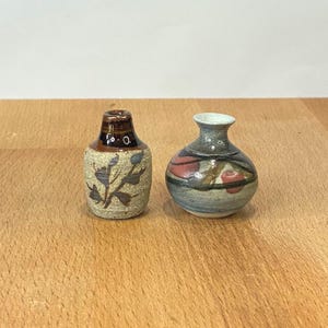 May include: Two miniature ceramic vases. The left vase is cylindrical with a speckled beige body, a dark brown neck, and a dark brown leaf pattern. The right vase is spherical with a flared rim, featuring a blue, grey, and red painted design.