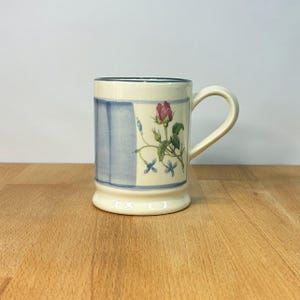 Może przedstawiać: Kremowy kubek ceramiczny z namalowanym wzorem różowego pąka róży z zielonymi liśćmi i małymi niebieskimi kwiatami. Kubek posiada pionowy wzór w niebieskie paski z boku oraz ciemnoniebieską obwódkę wzdłuż górnej krawędzi z uchwytem.