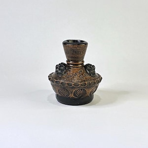 May include: A small, dark brown ceramic candle holder with a wide, flared neck and a rounded body. The surface features embossed circular patterns and leaf motifs, with two small, dark, sculpted animal-like figures attached to the shoulder.