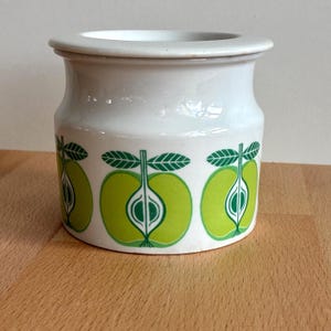 May include: A white ceramic jar with a lid, decorated with a repeating pattern of green apple halves. The apples have green leaves and stems. The jar sits on a wooden surface.