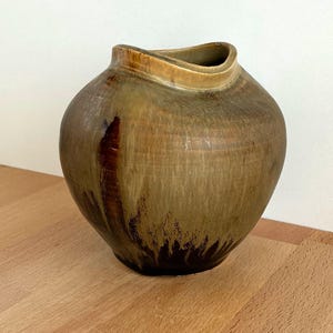 Puede incluir: Un jarrón de cerámica hecho a mano con una forma orgánica única. El jarrón presenta una mezcla de tonos terrosos, incluyendo verde oliva, marrón y beige, con una superficie texturizada. La abertura tiene un diseño asimétrico distintivo, lo que aumenta su atractivo artístico. El jarrón está sobre una superficie de madera.