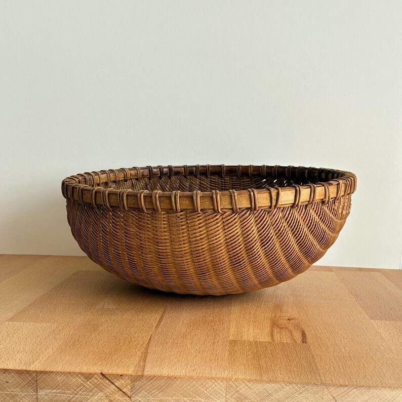 Pu&ograve; includere: Un cesto rotondo intrecciato di colore marrone naturale. Il cesto ha un design ampio e aperto e un bordo robusto. Il motivo di tessitura crea una superficie strutturata. Il cesto &egrave; appoggiato su una superficie di legno.