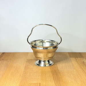 Puede incluir: Una pequeña cesta de metal plateado pulido con un cuenco redondeado, una base de pedestal acampanada y un asa fina y arqueada. El interior del cuenco es liso y reflectante, mientras que el exterior presenta sutiles estrías horizontales.