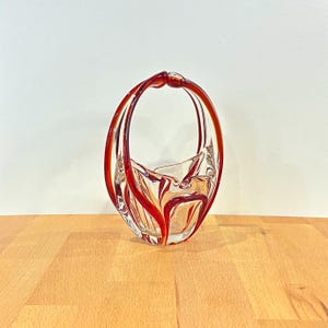May include: A clear glass basket with a red handle and accents. The basket has a unique, curved design with a handle that arches over the top. The red accents create a striking contrast against the clear glass, adding visual interest to the decorative piece.