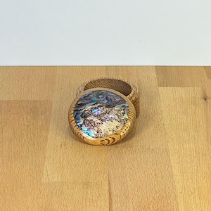 Könnte beinhalten: Eine kleine, runde Holzschatulle mit einem Deckel, der mit einer eingelegten, irisierenden Abalone-Muschelscheibe verziert ist. Die Schatulle besteht aus hellem Holz mit sichtbarer Maserung, und der Deckel ist kreisförmig mit einer glatten Oberfläche.