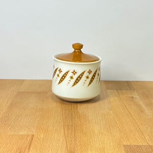 May include: A ceramic sugar bowl with a glossy off-white base and a solid brown lid. The exterior features a repeating pattern of brown diamond shapes containing small yellow and orange floral motifs arranged in a vertical, rhythmic sequence.