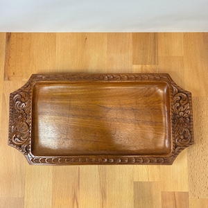 Puede incluir: Una bandeja rectangular de madera con un acabado liso y pulido y un centro hundido. Los dos extremos más cortos presentan intrincados patrones florales y de volutas tallados a mano, mientras que los bordes están bordeados por un motivo de hojas.