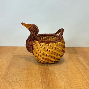 May include: A woven basket shaped like a duck, featuring a round body made of light tan reeds and a dark brown neck and head with a smooth, carved wooden beak. The basket has an open top with a woven rim and a small handle at the back.