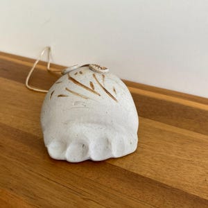 May include: A handmade, off-white ceramic bell with brown painted details and a natural fiber string. The bell has a rounded shape with a textured surface and small decorative elements on top. The bell is sitting on a wooden surface.