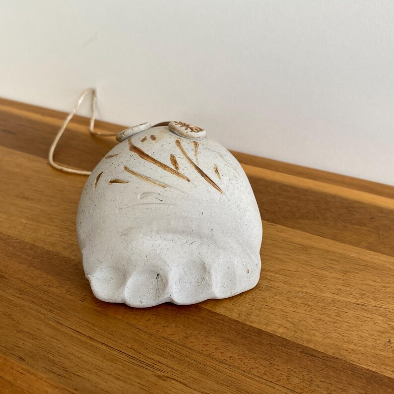May include: A handmade, off-white ceramic bell with brown painted details and a natural fiber string. The bell has a rounded shape with a textured surface and small decorative elements on top. The bell is sitting on a wooden surface.