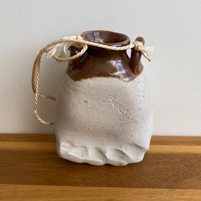 May include: A handmade ceramic jug with a brown top and a cream-colored body. The jug has a small handle and a rope tied around the neck. The word "Seer" is inscribed on the body of the jug.