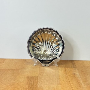 May include: A silver-toned metal dish shaped like a scallop shell with a beaded edge. The interior features a reflective, polished surface with embossed radial ridges and three small circular indentations, supported by a clear plastic stand.