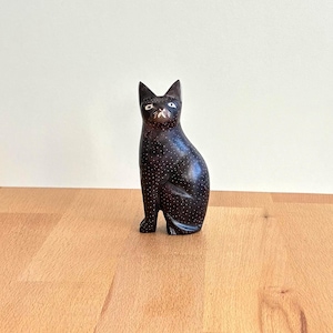 May include: A dark brown wooden cat figurine with white dot detailing. The cat is in a seated position with its ears perked up. The figurine is on a wooden surface.