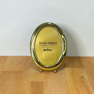 May include: An oval solid brass picture frame with a polished lacquer finish. The center insert features the text SOLID BRASS, hand polished & lacquer coated, 3 1/2" x 5" (9 cm x 13 cm), by Bowon. The frame stands on two small rounded brass feet.