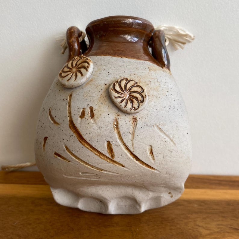 May include: A handmade ceramic vase with a speckled white body and a brown rim and handles. The vase features two decorative spiral accents and etched leaf-like patterns. A natural fiber cord is attached.