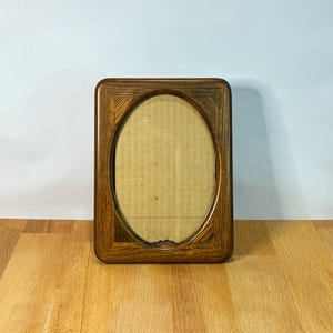Op de afbeelding: Een rechthoekige houten fotolijst met een donkerbruine afwerking en een opvallend houtnerfpatroon. De lijst heeft een centrale ovale opening die een eenvoudige, lichtbruine kartonnen achterkant in de lijst onthult.