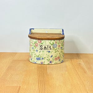 May include: A ceramic wall-mounted salt box with a cream-colored base featuring a floral pattern of small red, blue, and green flowers. The container has a hinged wooden lid and the word SALT printed in a rectangular black-outlined box on the front.