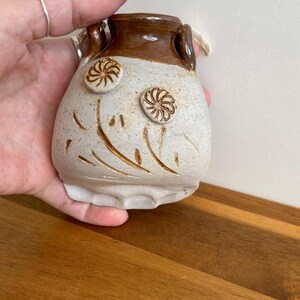 May include: A small, handmade ceramic vase with a speckled, off-white body and a brown rim. The vase features two raised, flower-like designs and etched floral patterns. It has two small handles and a textured, uneven base.