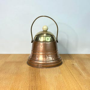 May include: A copper-coloured metal container with a wide, flared base and a rounded body. It features a polished brass lid topped with a small, cream-coloured spherical knob and a thin, arched metal handle attached to the sides of the vessel.