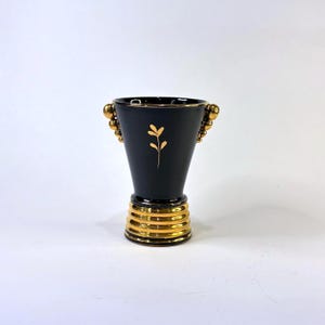 May include: A matte black ceramic vase with a flared rim and a tapered body. The vase features a gold-coloured floral sprig design on the front, gold-coloured beaded handles on both sides, and a ribbed gold-coloured base with horizontal rings.