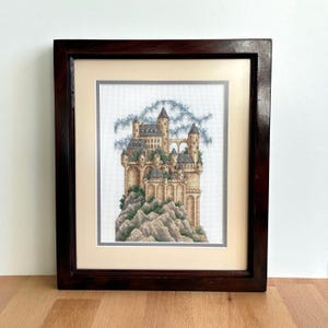 May include: Framed cross-stitch art depicting a detailed castle scene. The castle, in shades of tan, gray, and green, sits atop a rocky mountain. The artwork is framed in a dark brown wooden frame with a cream-colored mat.